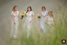 At St Peter & St Paul Church, Wing, Rutland, bridesmaids walk together along a dirt path bordered by tall green grass blades, the natural setting lending a fresh, serene backdrop to their procession.
